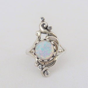 Pode incluir: Um anel de prata com uma pedra de opala branca e pequenos acentos de zircônia cúbica transparente. O anel tem um design delicado e ornamentado.