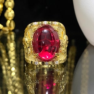 Puede incluir: Un anillo de oro con una gran piedra preciosa roja de forma ovalada engastada en el centro. El anillo está decorado con detalles intrincados y pequeñas piedras preciosas transparentes alrededor del borde.