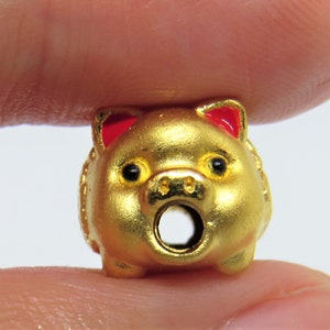 May include: A gold-colored pig charm with red ears. The pig has a round body and a hole through the center for attaching to a necklace or bracelet.