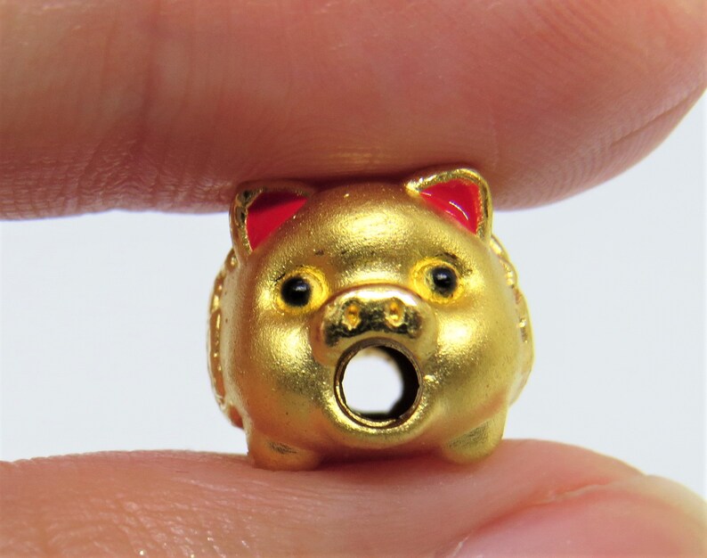 May include: A gold-colored pig charm with red ears. The pig has a round body and a hole through the center for attaching to a necklace or bracelet.