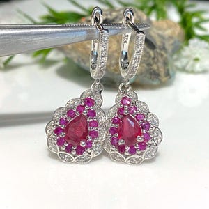 May include: A pair of silver dangle earrings with a pear-shaped ruby gemstone surrounded by smaller ruby and clear stones. The earrings have a hoop design with a 925 stamp.