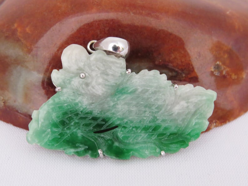 Pode incluir: Um pingente de jade verde e branco esculpido em forma de folha com detalhes prateados.