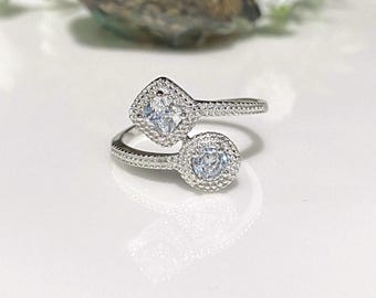 May include: A silver-toned ring with two clear, sparkling stones. One stone is square, the other is round, both surrounded by smaller, clear stones. The ring's band is also detailed with small, clear stones, creating a dazzling effect. The ring is set against a white background.