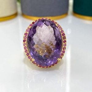 May include: A large, oval-shaped amethyst ring with a gold band. The amethyst is surrounded by a halo of small, round, pink gemstones. The ring is set against a white surface.