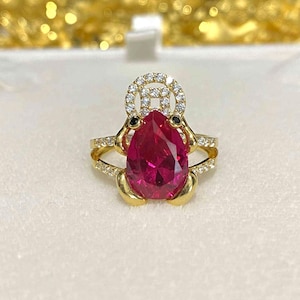 May include: A gold ring featuring a large, pear-shaped, ruby-red gemstone. The ring is accented with smaller, clear gemstones and black accents. The ring has a unique design with a halo of clear gemstones above the ruby.