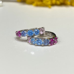 May include: A pair of silver hoop earrings with a row of colorful gemstones. The earrings are set with pink, blue, and light blue stones.