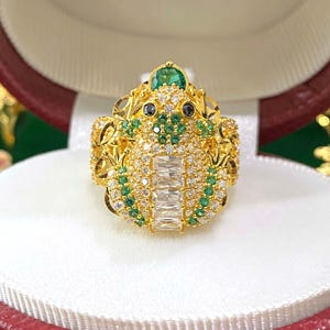 以下が含まれることがあります： カエルのデザインの金の指輪。カエルは緑と白の宝石で飾られています。指輪は赤と白の箱に入っています。
