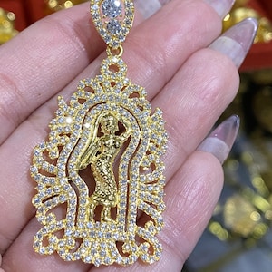 May include: A gold-colored pendant with a detailed design featuring a central figure and encrusted with clear gemstones. The pendant has a teardrop-shaped top with two larger stones and a decorative frame.