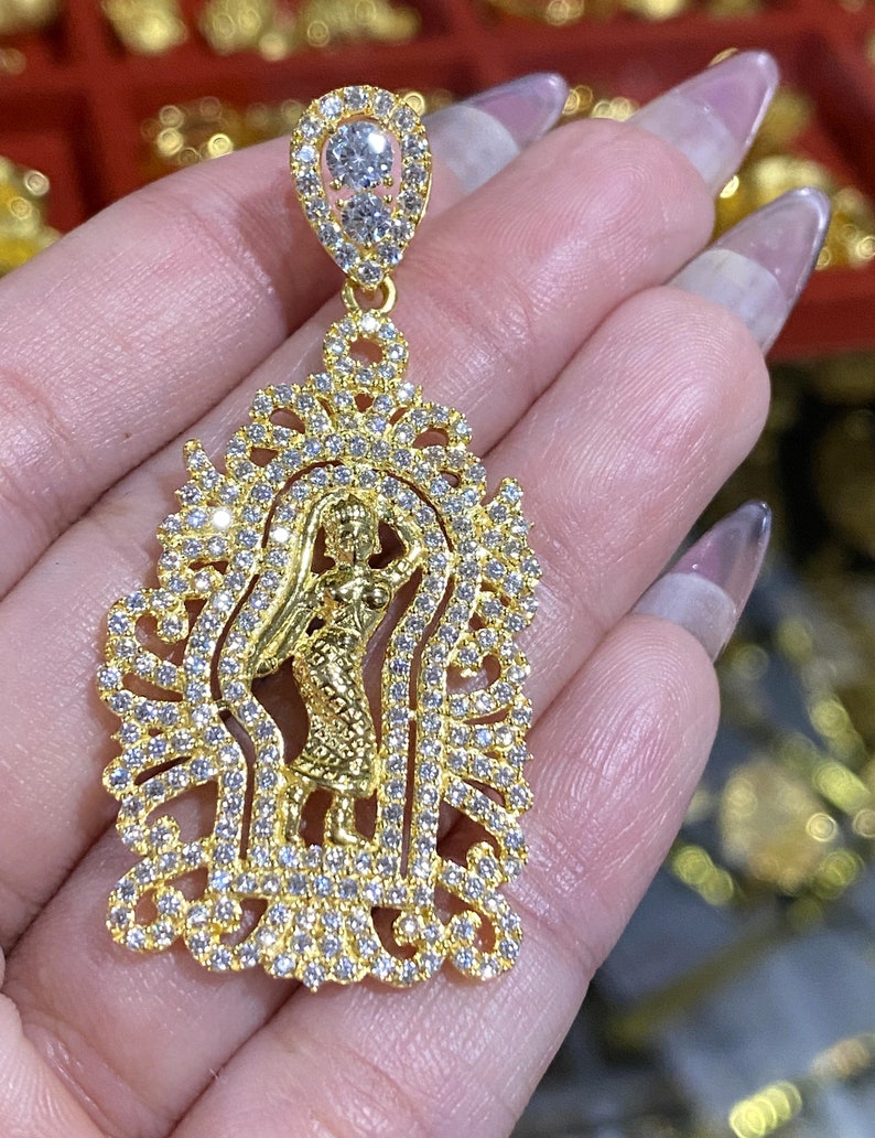 May include: A gold-colored pendant with a detailed design featuring a central figure and encrusted with clear gemstones. The pendant has a teardrop-shaped top with two larger stones and a decorative frame.