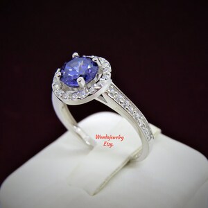 May include: A silver ring with a large, round, purple gemstone surrounded by a halo of smaller, white gemstones. The band is also set with smaller, white gemstones.