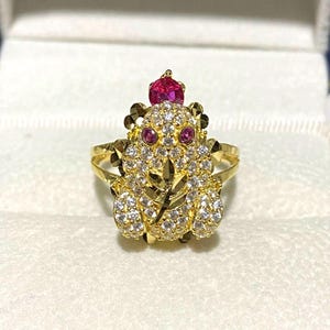 Peut inclure: Bague dorée représentant une grenouille ornée de pierres précieuses claires et rouges. Le corps de la grenouille est incrusté de pierres claires, avec des pierres rouges pour les yeux et une pierre rouge plus grande sur la tête. Sur fond blanc.
