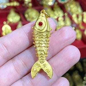 Puede incluir: Un amuleto de pez de color dorado con un ojo de piedra roja. El amuleto tiene escamas detalladas y caracteres chinos en la cola.
