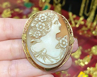May include: A gold-toned cameo brooch with a detailed carving of a woman's profile surrounded by flowers. The cameo is set in a delicate filigree frame.