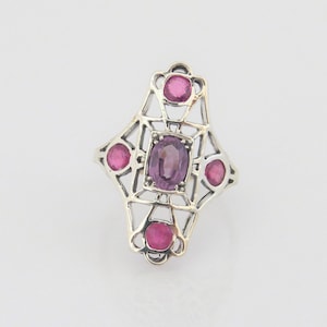 May include: A silver ring with a geometric design featuring a large oval amethyst gemstone surrounded by four smaller round ruby gemstones.