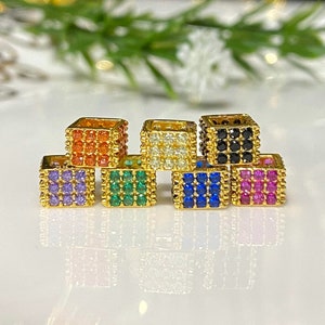 May include: A set of six gold-toned charm pendants, each featuring a square shape with a different colored crystal inlay. The charms are arranged in a row, with the colors from left to right being purple, green, white, black, blue, and pink.