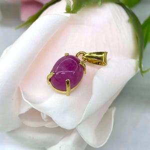 Puede incluir: Un colgante de oro con una piedra preciosa ovalada rosa engastada en un engaste de oro. El colgante est&aacute; sobre un fondo blanco.
