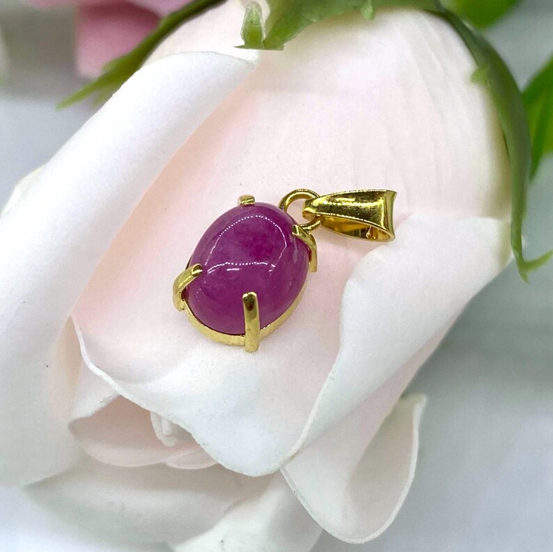 Puede incluir: Un colgante de oro con una piedra preciosa ovalada rosa engastada en un engaste de oro. El colgante est&aacute; sobre un fondo blanco.