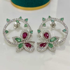 May include: A pair of silver butterfly earrings. Each earring features a butterfly design with ruby and emerald gemstones, surrounded by a halo of clear crystals. The earrings have a post back closure and a delicate, elegant appearance.