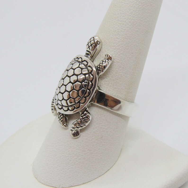 May include: A silver turtle ring with a detailed textured shell and legs. The ring is on a white display stand.