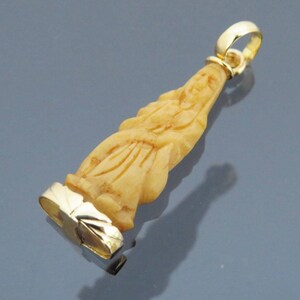 May include: A pendant featuring a carved figure in a light beige hue, with a gold-colored bail and a gold-colored base. The pendant is elongated and detailed, with visible carving lines. The background is a gradient of gray and blue.
