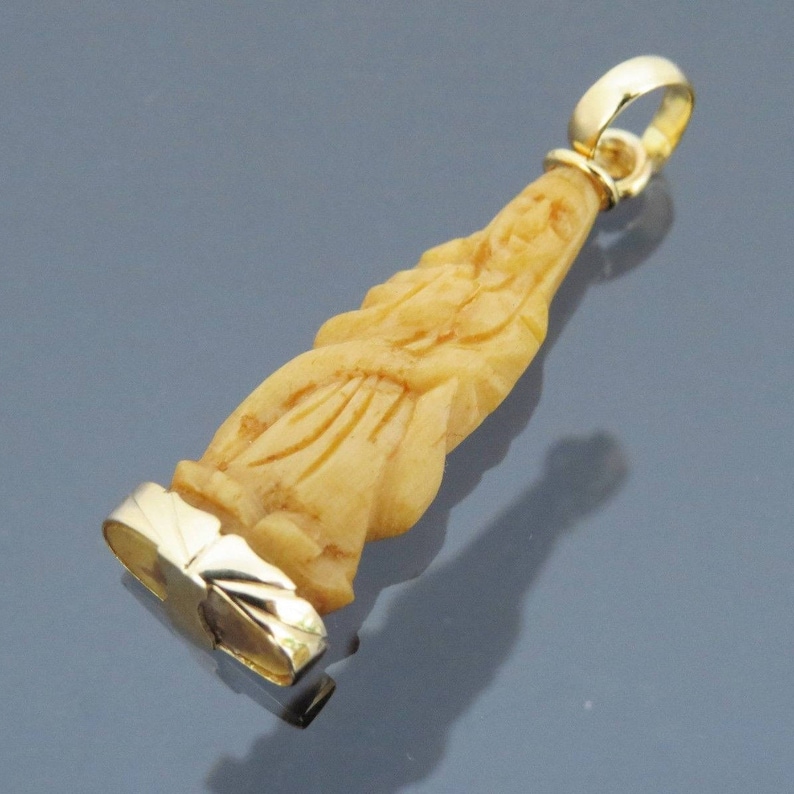 May include: A pendant featuring a carved figure in a light beige hue, with a gold-colored bail and a gold-colored base. The pendant is elongated and detailed, with visible carving lines. The background is a gradient of gray and blue.