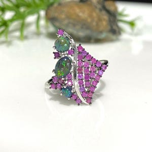 May include: A silver ring featuring a cluster of oval-shaped, iridescent black opals and a fan-shaped arrangement of small, round, pink gemstones. The ring's design is set against a white background.