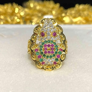 May include: A gold-toned ring with an ornate design. The ring features a central cluster of green, pink, and white gemstones, with red gemstones as eyes. The ring is set on a white surface.