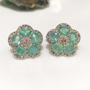 May include: A pair of flower-shaped stud earrings. Each earring features six oval-cut, light green emeralds surrounded by small pink, yellow, and clear gemstones. The earrings have a silver-colored metal setting.