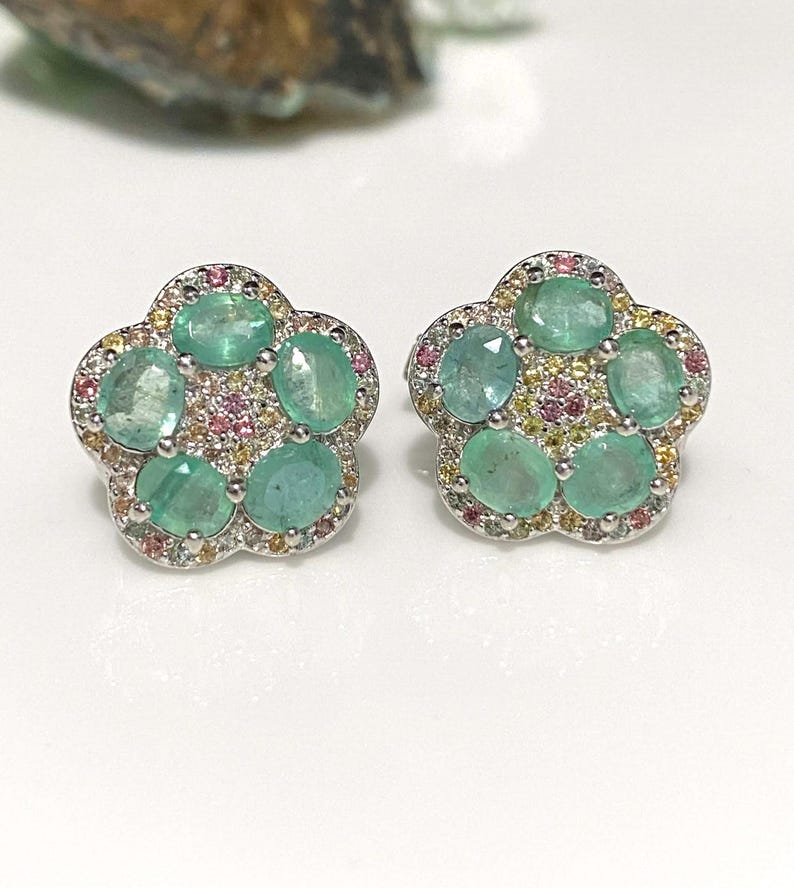 May include: A pair of flower-shaped stud earrings. Each earring features six oval-cut, light green emeralds surrounded by small pink, yellow, and clear gemstones. The earrings have a silver-colored metal setting.