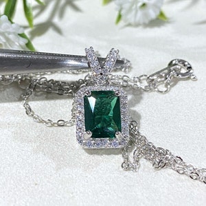 Könnte beinhalten: Eine silberne Halskette mit einem grünen Smaragd-Anhänger, der von kleineren klaren Steinen umgeben ist. Der Anhänger hat eine rechteckige Form mit einer Halo-Fassung.