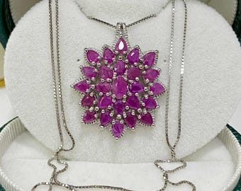 May include: A silver necklace with a pendant of deep pink gemstones in an oval cut, arranged in a floral design. The necklace is presented in a green and white jewellery box.