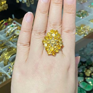 May include: A gold ring with a cluster of yellow gemstones. The ring is worn on the index finger of a hand.