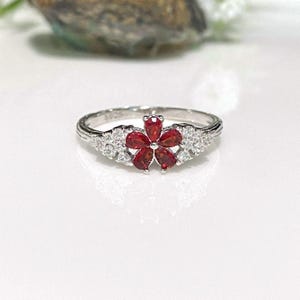 May include: A silver ring featuring a flower design with red gemstones at the center and small white gemstones on the sides. The ring is set on a white surface.