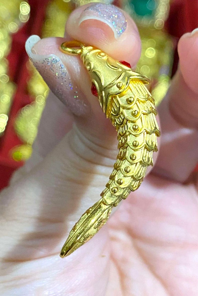 Puede incluir: Un amuleto de pez de color dorado con un ojo de piedra roja. El amuleto est&aacute; detallado con escamas y tiene un bucle para sujetarlo a un collar o pulsera.