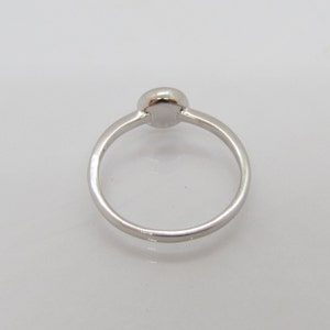 Puede incluir: Un anillo plateado con una piedra central redonda y elevada. La banda es fina y lisa, sobre un fondo blanco. El anillo es una joya sencilla y elegante.