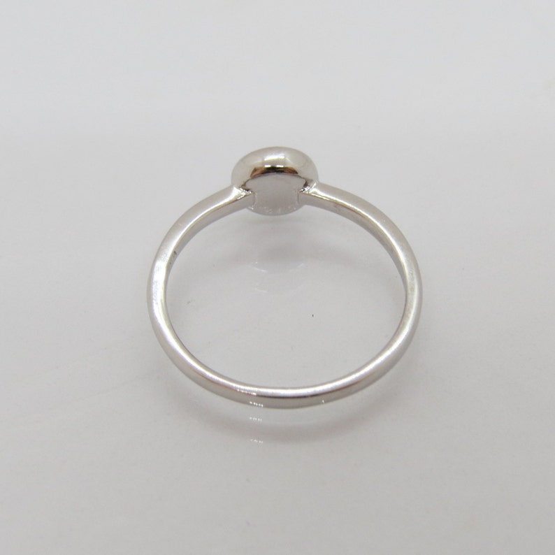 Puede incluir: Un anillo plateado con una piedra central redonda y elevada. La banda es fina y lisa, sobre un fondo blanco. El anillo es una joya sencilla y elegante.