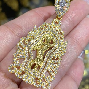 May include: A gold pendant featuring a detailed design with a central figure and an ornate, sparkling frame. The pendant is encrusted with numerous small, clear gemstones, and a larger gemstone is at the top.