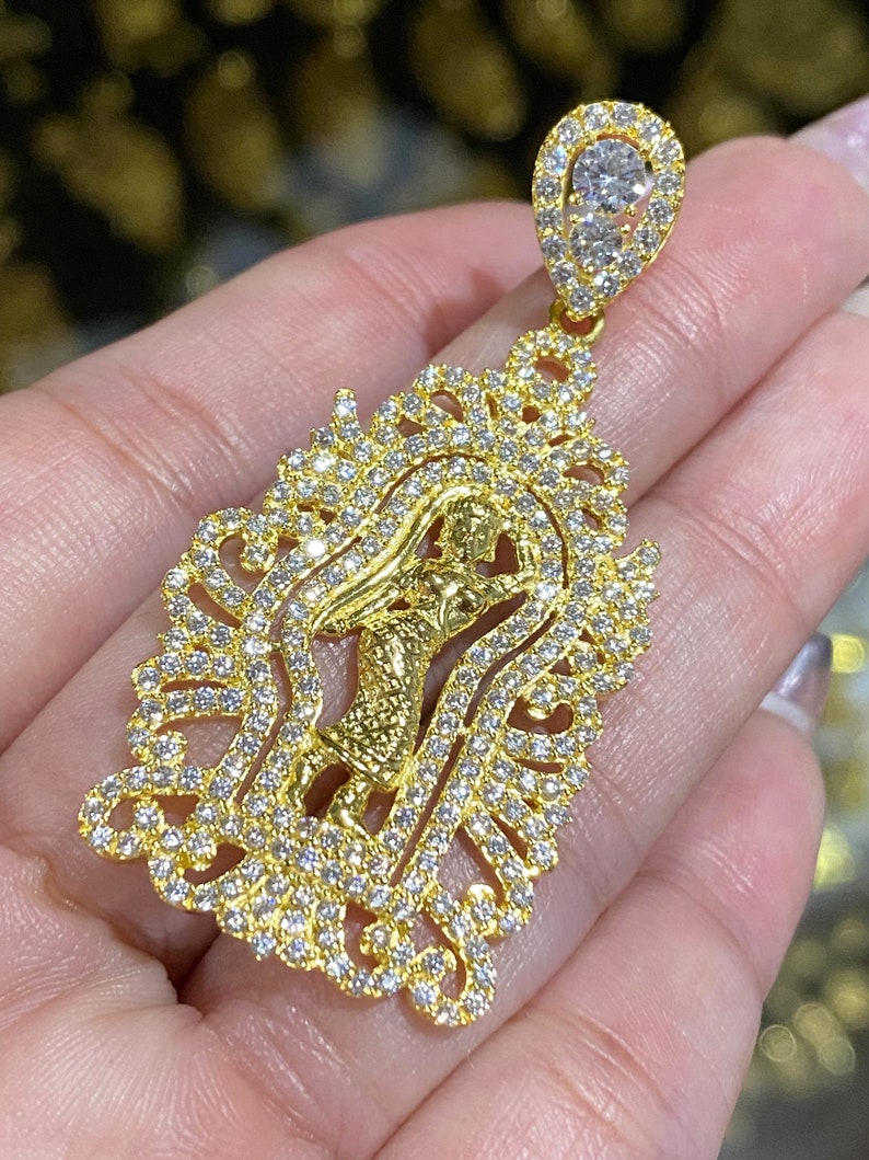 May include: A gold pendant featuring a detailed design with a central figure and an ornate, sparkling frame. The pendant is encrusted with numerous small, clear gemstones, and a larger gemstone is at the top.
