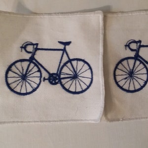 Può includere: Due sottobicchieri quadrati bianchi con un disegno di bicicletta blu navy. La bicicletta è in stile classico, con ruote e telaio dettagliati. I sottobicchieri sono realizzati in un tessuto morbido e strutturato.