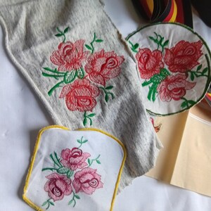 May include: Three fabric pieces featuring floral embroidery. One is a heather gray rectangular strip with red roses, one is a circular white patch with red roses and green stems, and one is a white square patch with pink roses and a yellow border.