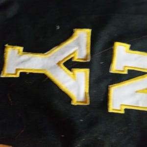 Peut inclure: Une surface en tissu noir présentant deux grandes lettres blanches de style bloc avec d'épaisses bordures brodées jaune vif. Les lettres sont positionnées horizontalement, le caractère de gauche ressemblant à un Y stylisé et celui de droite à un Z.