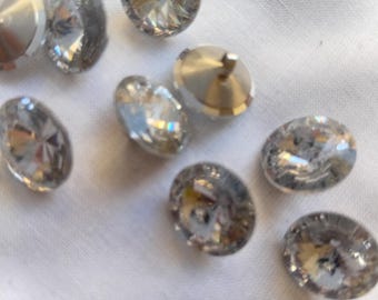 Könnte beinhalten: Nahaufnahme mehrerer klarer, facettierter Kristallverzierungen. Die Kristalle sind oval und rund, mit einer metallischen Rückseite. Einige haben einen Stift zum Befestigen an Stoff oder anderen Materialien. Die Kristalle sind auf einer weißen Oberfläche verstreut.