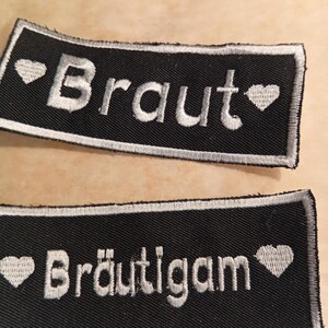 Op de afbeelding: Twee zwarte rechthoekige patches met wit geborduurde tekst en hartsymbolen. Eén patch leest "Braut" en de andere "Bräutigam". Beide patches hebben een witte rand.