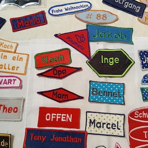 Peut inclure: Une collection d'écussons en tissu brodé de formes et de couleurs variées, présentant des noms et des mots tels que Sydney, Maria, Frohe Weihnachten, # 88, Jakob, Piepmatz, Noah, Opa, Inge, Mam, Bennet, OFFEN, Marcel, Theo, Tony Jonathan, Danny et Vivian.