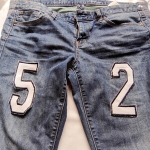 Könnte beinhalten: Blaue Jeans aus Denim mit einem weißen Stoffaufnäher der Zahl 5 auf dem linken Oberschenkel und der Zahl 2 auf dem rechten Oberschenkel. Die Hose verfügt über ein klassisches Fünf-Taschen-Design, einen Knopfverschluss und ein Tommy Hilfiger Etikett.