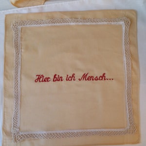 May include: A beige handkerchief with a white lace border. The phrase "Hier bin ich Mensch..." is embroidered in red across the centre. The handkerchief is rectangular and lies on a white surface.