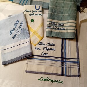 May include: A collection of embroidered handkerchiefs in various colors, including blue, green, and yellow. Some handkerchiefs feature text such as "Alles Gute zum Geburtstag" and "Alles Liebe zum 60 igsten." The handkerchiefs have striped and patterned borders.