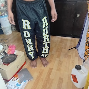May include: Black athletic trousers featuring a yellow and white block letter graphic running vertically down each leg. The left leg displays the text RONNY and the right leg displays the text HURTHDE. The trousers have an elastic waistband.
