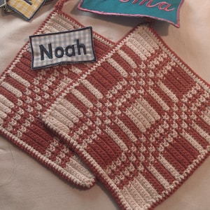 Peut inclure: Deux maniques faites à la main, rouges et crème, avec un motif à carreaux. Un petit patch avec le nom "Noah" en broderie bleue est attaché. D'autres patchs de noms sont visibles en arrière-plan.