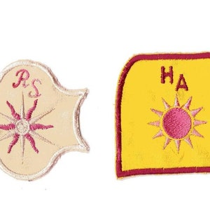 Peut inclure: Deux écussons en tissu brodé. L'écusson de gauche est de couleur crème avec un bord festonné, présentant un motif de soleil rose et les lettres RS. L'écusson de droite est jaune avec une bordure rose foncé, un soleil rose et les lettres HA.